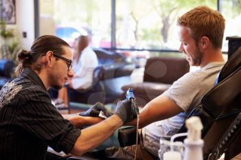 Man Sitting In Chair Having Tattoo On Arm In Parlor
