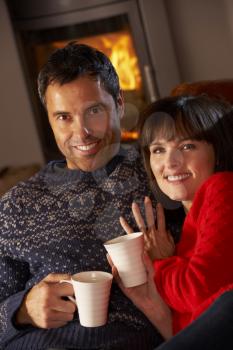 Middle Aged Couple Sitting On Sofa By Cosy Log Fire With Hot Drink