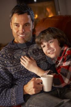 Father And Son Relaxing With Hot Drink Watching TV By Cosy Log Fire