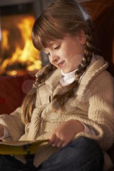 Young Girl Sitting On Sofa And Reading Book By Cosy Log Fire