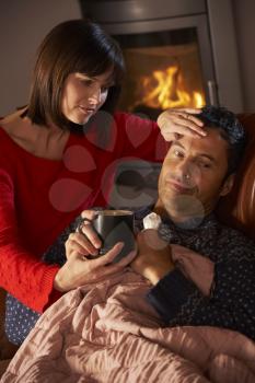 Wife Nursing Sick Husband With Cold Resting On Sofa By Cosy Log Fire