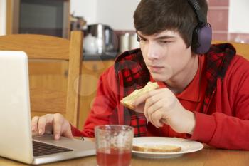 Teenage Boy Using Laptop Whilst Eating Breakfast