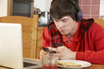 Teenage Boy Using Laptop And Listening To MP3 Player Whilst Eating Breakfast