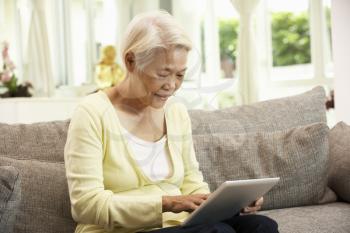 Senior Chinese Woman With Tablet Computer Whilst Relaxing On Sofa At Home