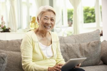 Senior Chinese Woman With Tablet Computer Whilst Relaxing On Sofa At Home