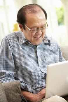 Senior Chinese Man Using Laptop Whilst Relaxing On Sofa At Home