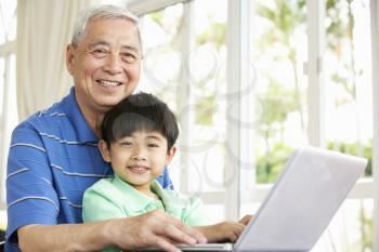 Chinese Grandfather And Grandson Sitting At Desk Using Laptop At Home