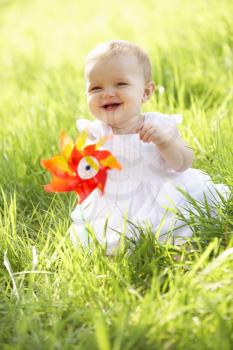 Baby Girl In Summer Dress Sitting In Field Holding Windmill
