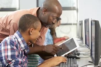 Teacher Helping Boy To Use Digital Tablet In Computer Class
