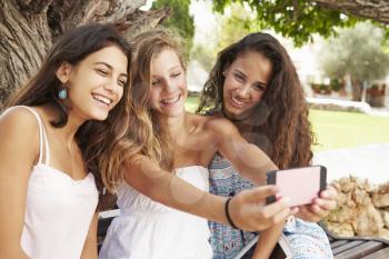 Three Teenage Girls Sitting On Bench Taking Selfie In Park