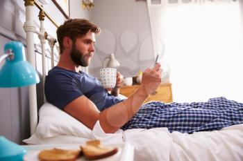 Man Eating Breakfast In Bed Whilst Using Mobile Phone