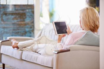 Mature Woman Relaxing On Sofa At Home Using Digital Tablet