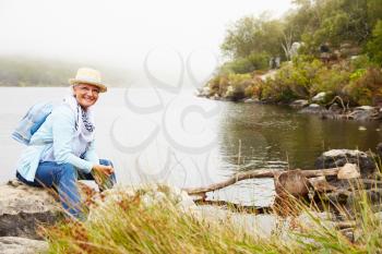 Senior woman with a hat sitting by a lake, smiling to camera