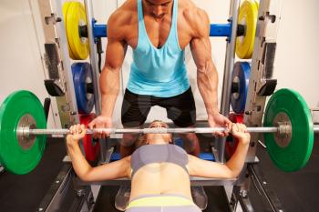 Woman bench pressing weights with assistance of trainer, front view
