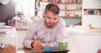 Young Man Eating Breakfast Whilst Using Digital Tablet