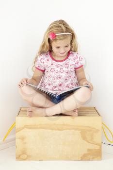 Young Girl Sitting On Wooden Toy Box Reading Book In Bedroom