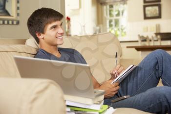 Teenage Boy Sitting On Sofa At Home Doing Homework Using Laptop Computer Whilst Watching TV
