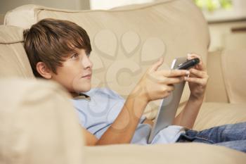Young Boy Sitting On Sofa At Home Using Tablet Computer Whilst Watching Television