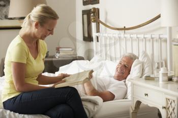 Adult Daughter Reading To Senior Male Parent In Bed At Home