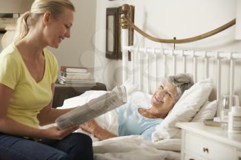 Adult Daughter Reading Newspaper To Senior Female Parent In Bed At Home