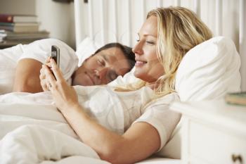 Woman Using Mobile Phone In Bed Whilst Husband Sleeps