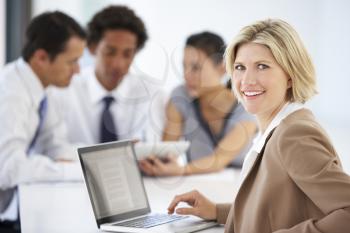 Portrait Of Female Executive Using Laptop With Office Meeting In Background