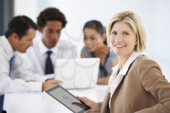 Portrait Of Female Executive Using Tablet Computer With Office Meeting In Background