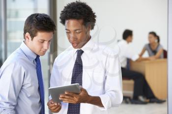 Two Male Executives Looking At Tablet Computer With Office Meeting In Background