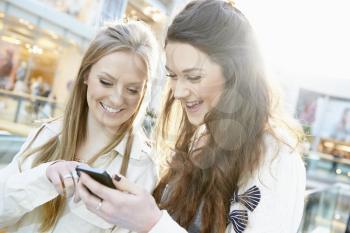 Two Female Friends Shopping In Mall Looking At Mobile Phone