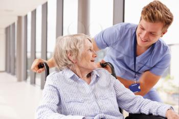 Nurse Pushing Senior Patient In Wheelchair Along Corridor