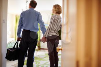 Senior couple walk in to hotel room holding hands, back view