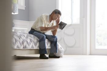 Unhappy Senior Man Sitting On Bed Looking At Photo Frame
