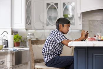 Young Asian Boy Playing Game On Mobile Device In Kitchen