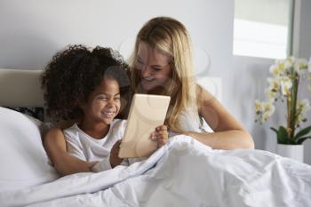 Black girl holds tablet computer, in bed with caucasian mum