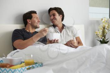 Male gay couple relax in bed eating, looking at each other