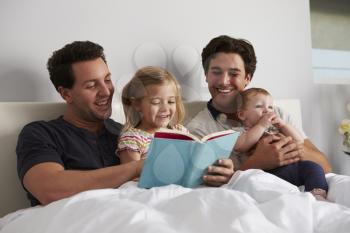 Male gay parents reading a book in bed with two kids