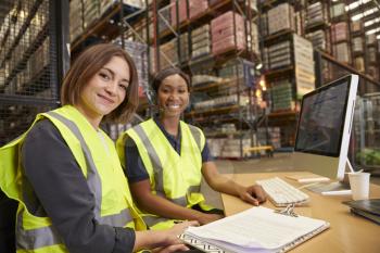 Two female colleagues in a warehouse office look to camera