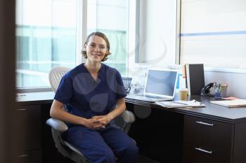 Portrait Of Nurse Wearing Scrubs Sitting At Desk In Office