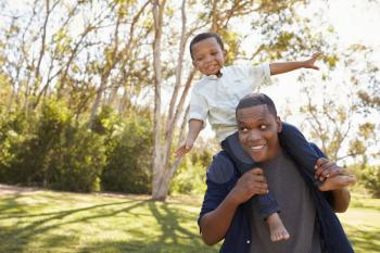 Father Carrying Son On Shoulders As They Walk In Park