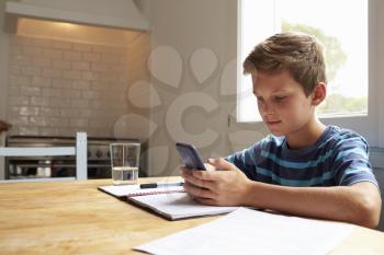 Boy Uses Mobile Phone Whilst Doing Homework At Kitchen Table