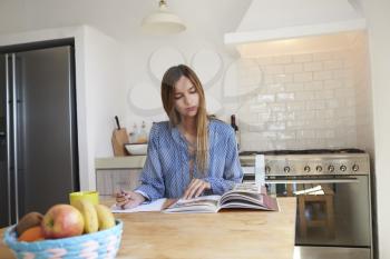 Young woman at kitchen table with recipe book writing a list