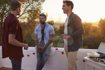 Three male friends talk at a party on a rooftop at sunset