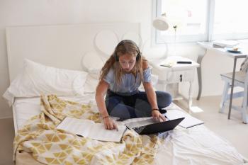 Teenage girl reading homework sitting on her bed with laptop