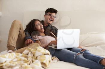 Laughing teenage couple hanging out in bedroom using laptop