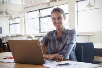 Young woman in office using laptop, smiling to camera