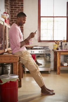 Young black man using smartphone in kitchen, full length