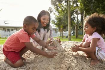 Teacher At Montessori School Playing With Children In Sand Pit