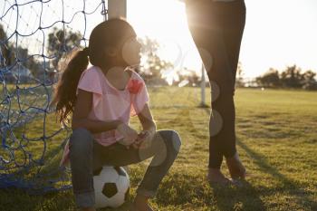 Young girl sits on ball next to her mum on a football pitch