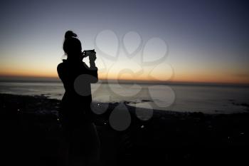 Woman taking photo with phone on beach, silhouette at sunset