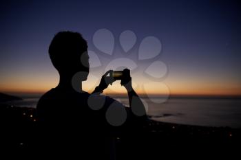 Man taking photos with phone on beach, silhouette at sunset